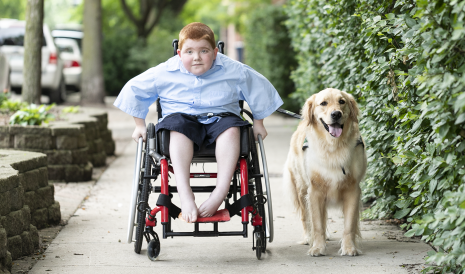 EXONDYS 51 patient Ryan and his dog EXONDYS 51 patient Ryan, age 11, seated in wheelchair on sidewalk rolling toward camera with his Golden Retriever by his side